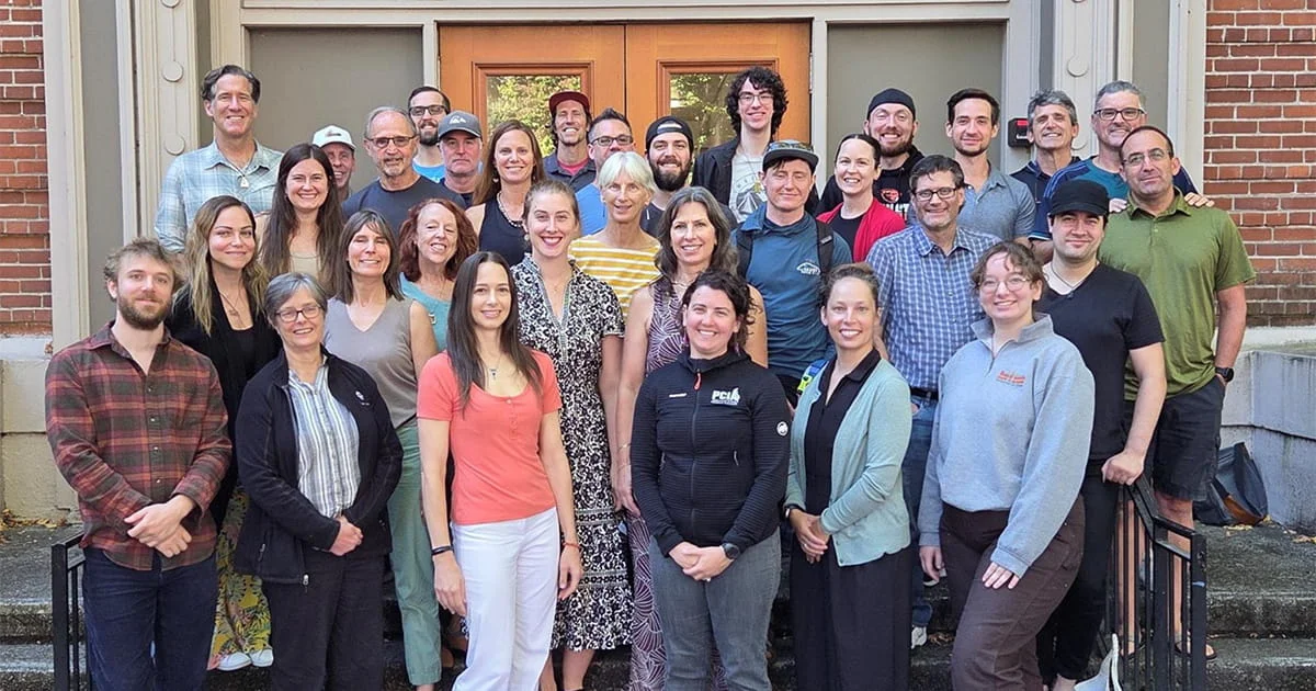 Group portrait of approximately 30 staff members standing and smiling on the steps in front of a brick building with large wooden doors.