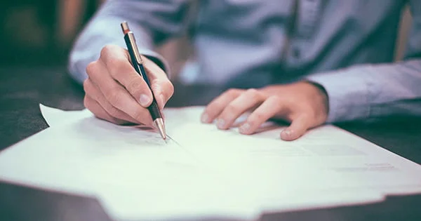 Close-up of a person's hand holding a pen and writing or signing a document or form on a desk, symbolizing paperwork completion or enrollment.
