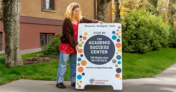 Student setting up a colorful, dot-patterned sidewalk sign for the Academic Success Center outside a brick campus building on a sunny day.