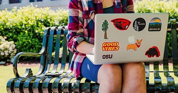 A student sits on a black metal bench on a sunny campus day, working on a laptop adorned with many personal stickers, including the Oregon State University beaver logo.