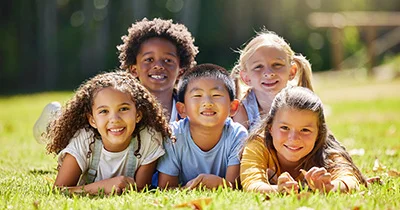 Five happy children of diverse backgrounds posing closely together on green grass during an outdoor activity on a sunny day.