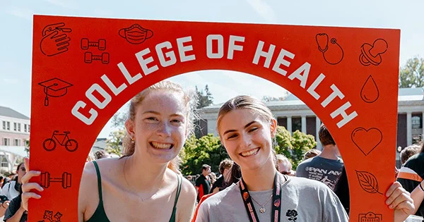 Two young women smiling in front of a large red banner that says "COLLEGE OF HEALTH".