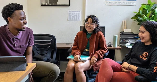 Three diverse college students sitting in an office, laughing and engaging in a casual but focused discussion, likely for a study group or lab.