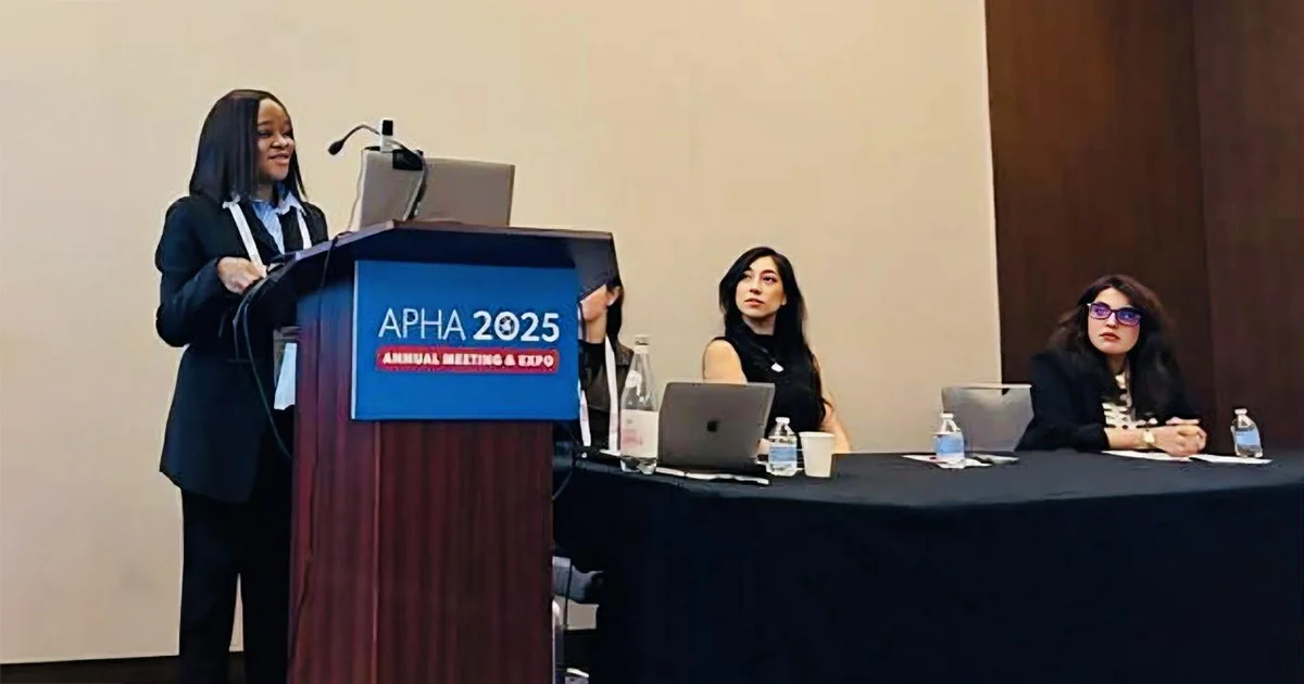 Speaker at a professional conference presenting from a podium labeled APHA 2025 to a panel of two seated attendees.