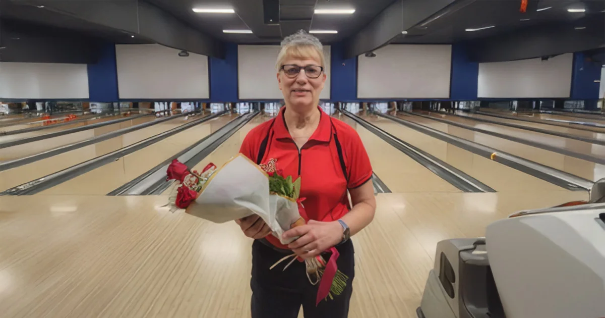 Smiling woman in a red shirt holding a bouquet of roses while standing in the middle of a bowling alley lane.