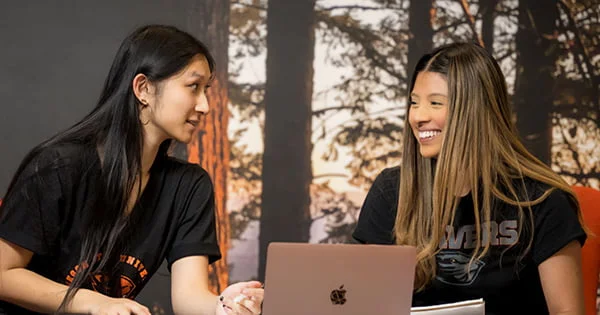 An in-hall advisor or staff member assists a student with her laptop in a residence hall common area, illustrating peer support services.