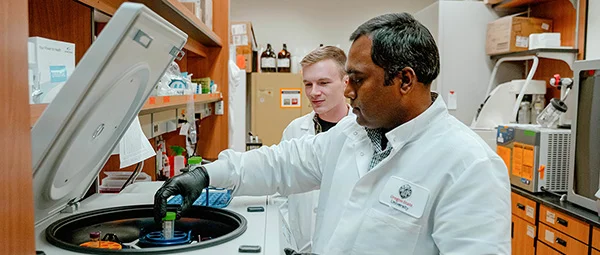 Two male researchers in lab coats are working with a centrifuge in a laboratory, with one researcher loading a sample tube into the machine.