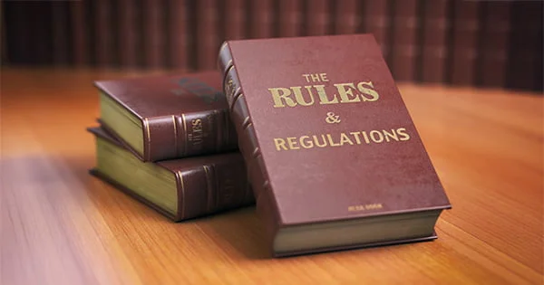 Close-up of three dark red hardcover books stacked on a wooden table. The front cover of the top book reads in gold letters: 'The Rules & Regulations'.