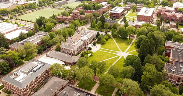 Drone photo of the Oregon State University campus on a sunny day, showcasing the mix of historic brick buildings and modern facilities amidst lush green lawns and abundant trees.