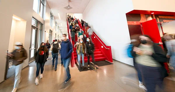 Students walking up and down a bright, modern red staircase and moving through a busy interior hallway on a university campus.