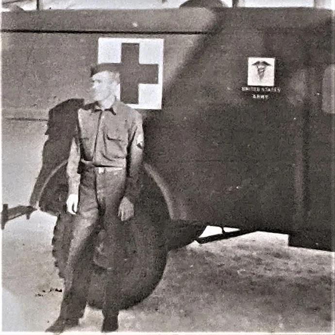 A vintage photograph of an Army soldier standing beside a military ambulance or medical transport vehicle.