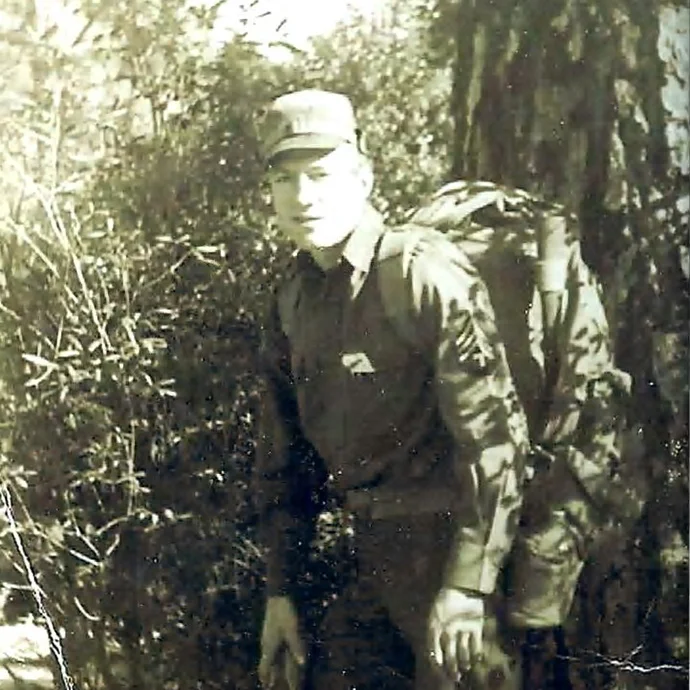 A vintage black and white photo of a smiling soldier wearing a large backpack in a wooded area.
