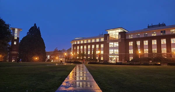 Exterior view of a large, modern university library at night. The windows are brightly lit, casting a warm glow onto a wet, reflecting sidewalk and surrounding grassy quad.