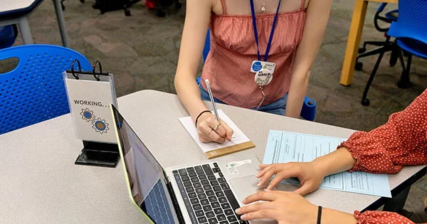 Collaborative session at a writing or learning center, showing students using both a laptop for digital work and a notepad for handwriting, indicating focused study and review.