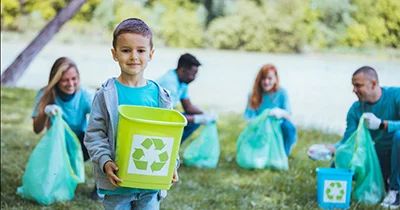 A young boy stands smiling in the foreground, holding a bright green recycling bin, while four adults in the background collect trash and recyclables in green plastic bags.