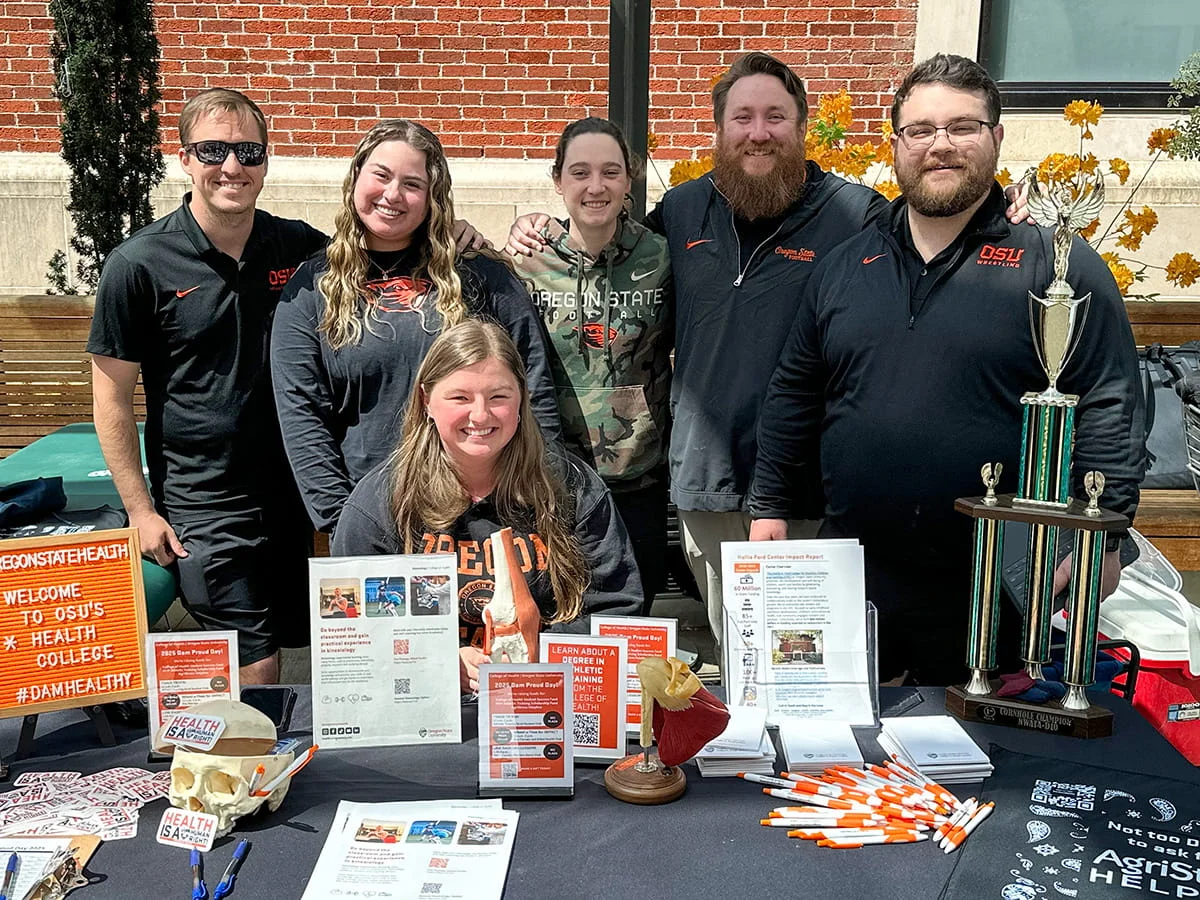 A group of Oregon State University students and staff pose at an outdoor health college resource fair. Their table displays educational materials and a cornhole championship trophy, illustrating campus engagement and school spirit.