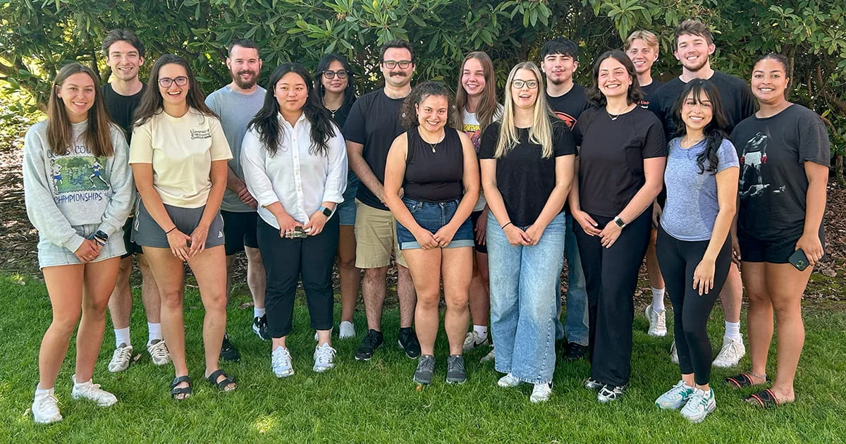 A group of fifteen diverse young adults stand in two rows, smiling and posing for a photo on a grassy lawn in front of dense green bushes.