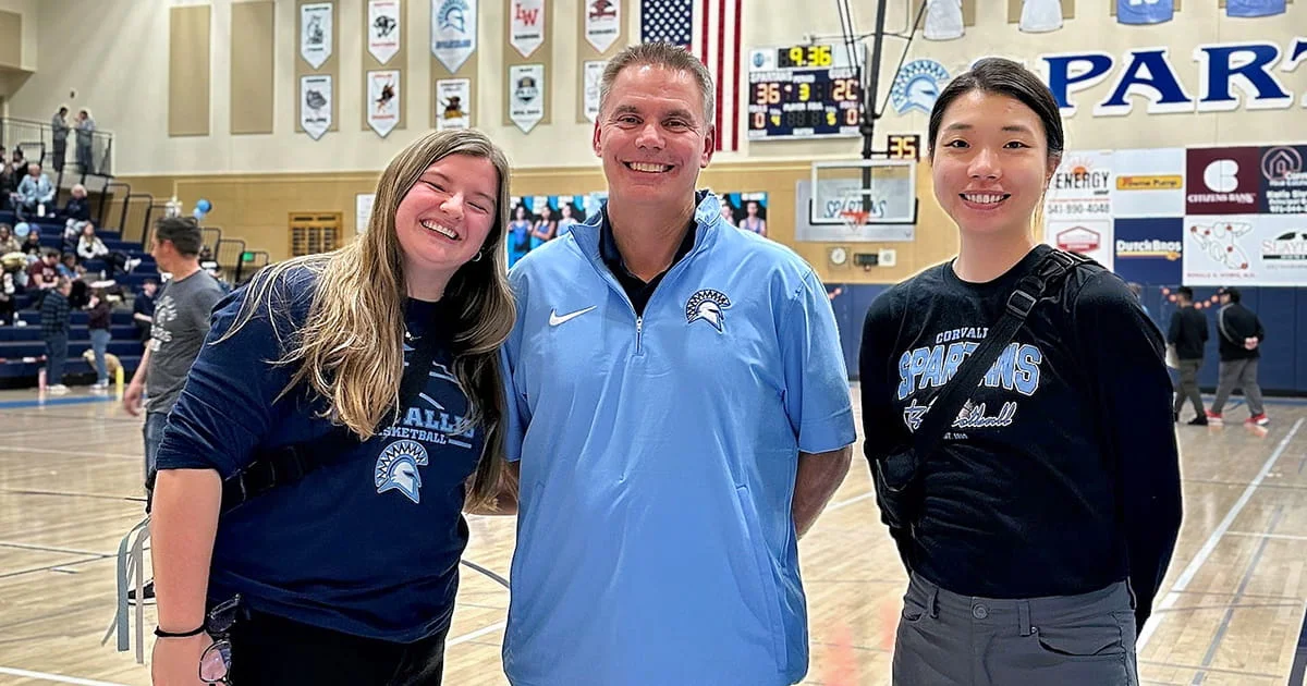A man in a light blue quarter-zip shirt stands between two young women wearing dark blue "Corvallis Spartans Basketball" shirts inside a high school gymnasium.