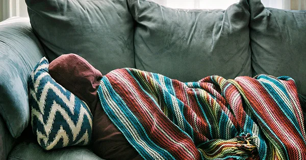 A person resting under a blanket on a couch, symbolizing comfort, shelter, and a need for housing or basic resources.