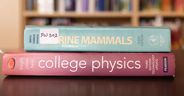 Textbooks for "College Physics" and "Marine Mammals," symbolizing academic study and the availability of resources through a textbook loan program.