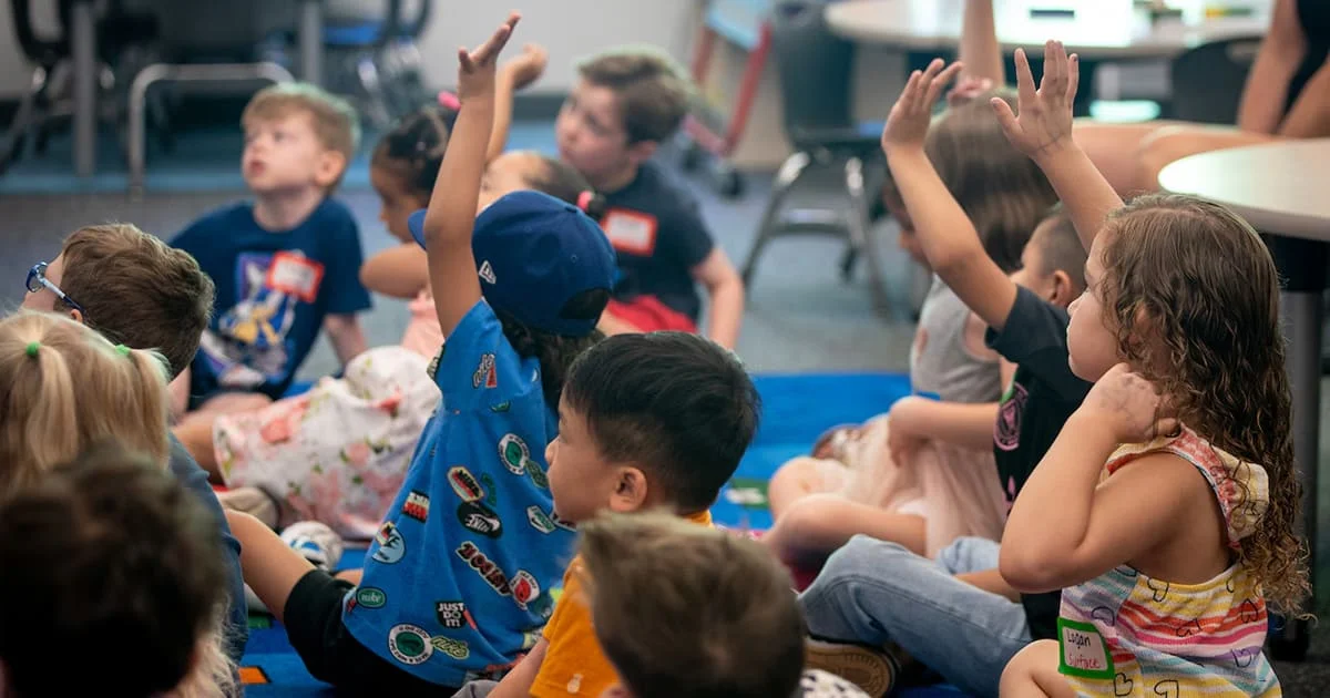 A group of young, diverse children sitting on a blue patterned carpet in a classroom, with several enthusiastically raising their hands to answer a question.