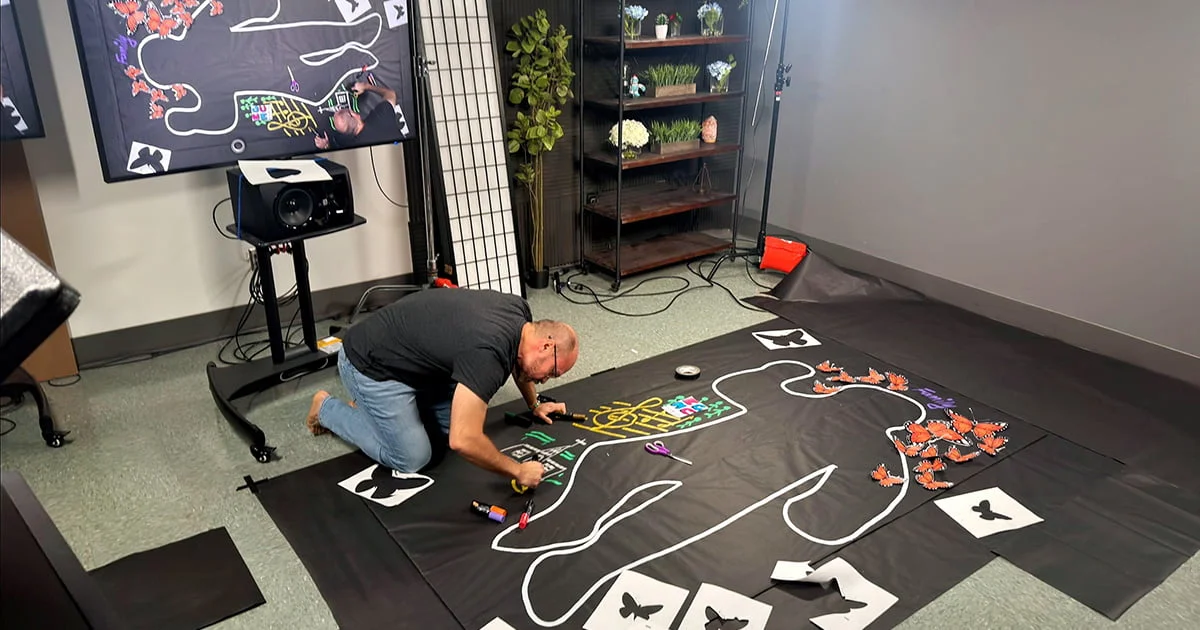 A man kneels on a studio floor, drawing a white chalk outline of a human figure on large black paper. Orange butterfly cutouts and various drawings decorate the installation.