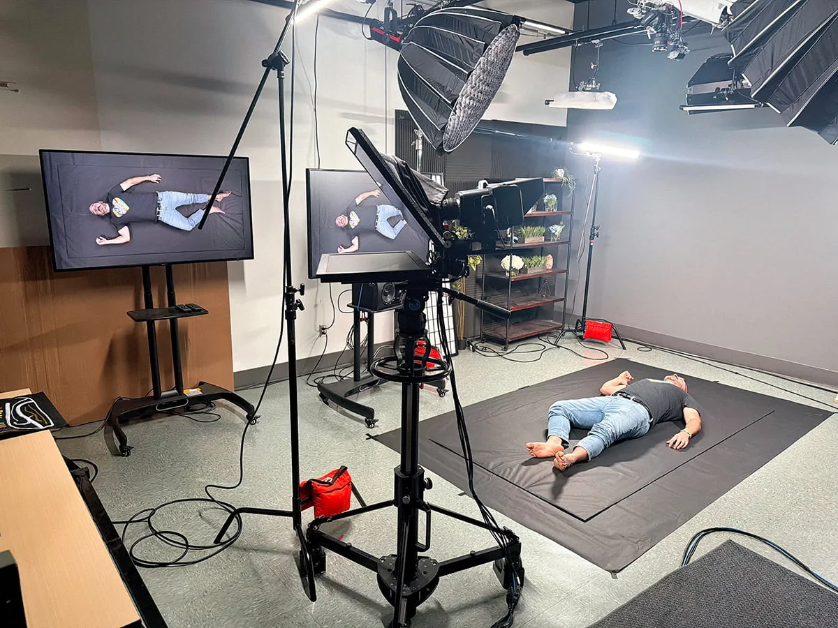 A man lies on his back on a large black mat in a professional production studio. A camera on a high tripod and several studio lights are positioned around him.