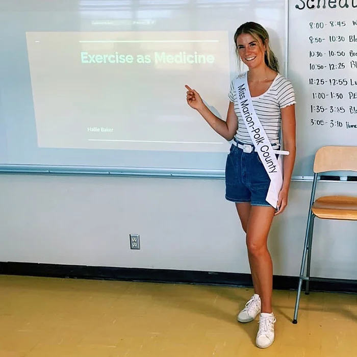 Hallie Baker, wearing a Miss Marion-Polk County sash, presents a slide titled "Exercise as Medicine" in a classroom.