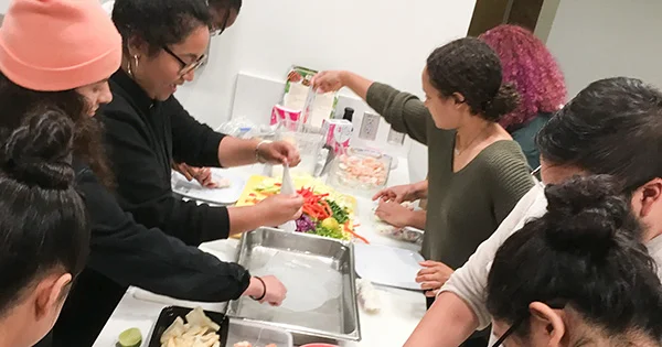 A group of diverse people gathered in a kitchen, collaboratively preparing food by rolling spring rolls and chopping colorful vegetables.
