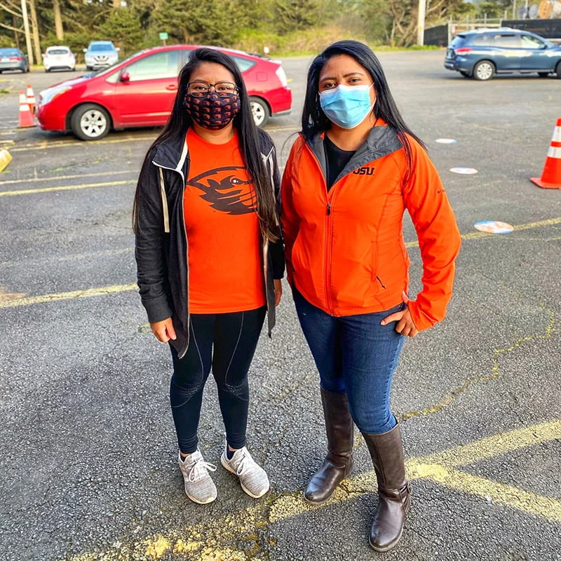 Two women wearing face masks stand side-by-side in a parking lot. One wears an orange shirt with a beaver logo; the other wears an orange OSU jacket.