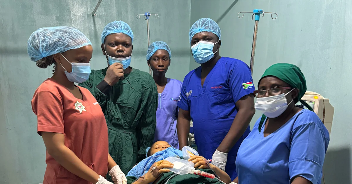 A medical team of five, wearing scrubs, surgical masks, and hairnets, stands around a patient lying on an operating table in a clinical setting.