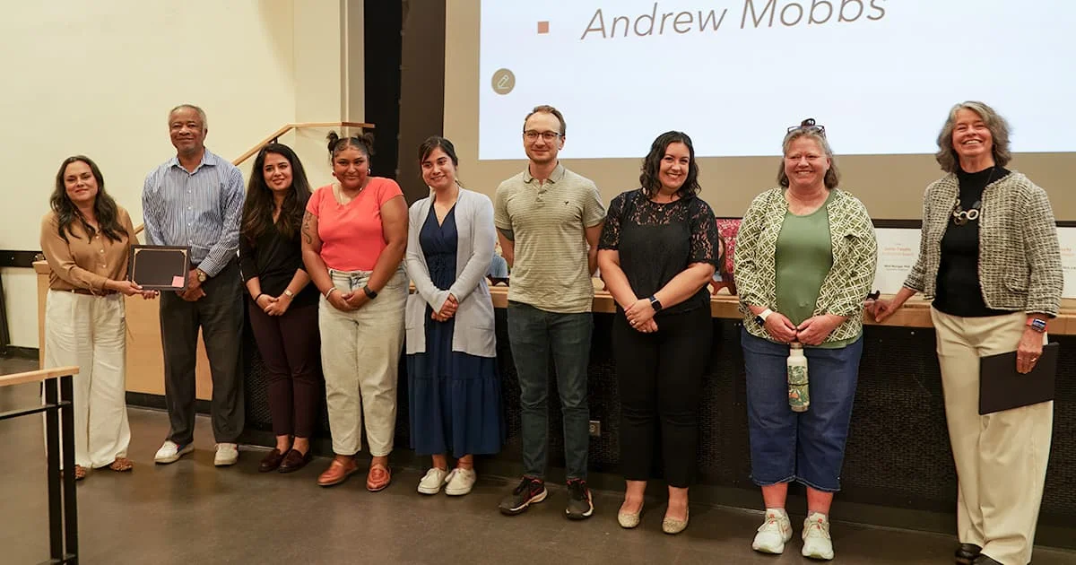 A group of nine diverse staff members stands in a row on a stage during an awards ceremony.
