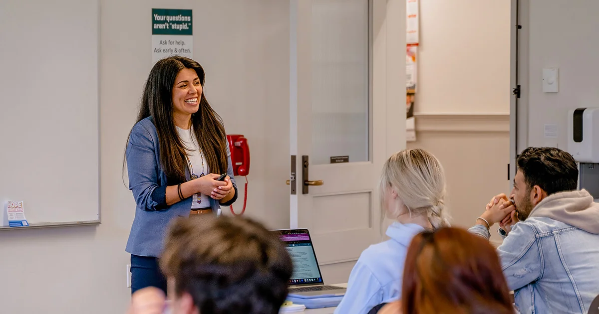 A supportive instructional setting at Oregon State University, where a smiling faculty member interacts with students. A wall poster in the background reads, "Your questions aren't 'stupid.' Ask for help. Ask early & often."