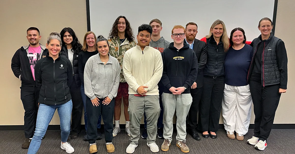 A diverse group of 14 smiling students and faculty members stand in two rows against a neutral wall in a professional meeting room setting.