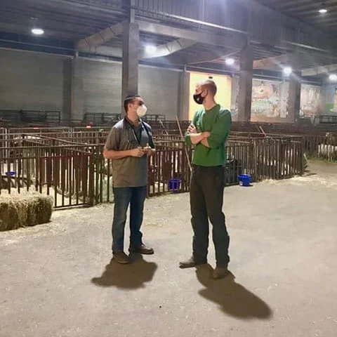 Two men wearing face masks stand in a large indoor livestock barn with animal pens in the background. One man wears a stethoscope around his neck while talking to the other.