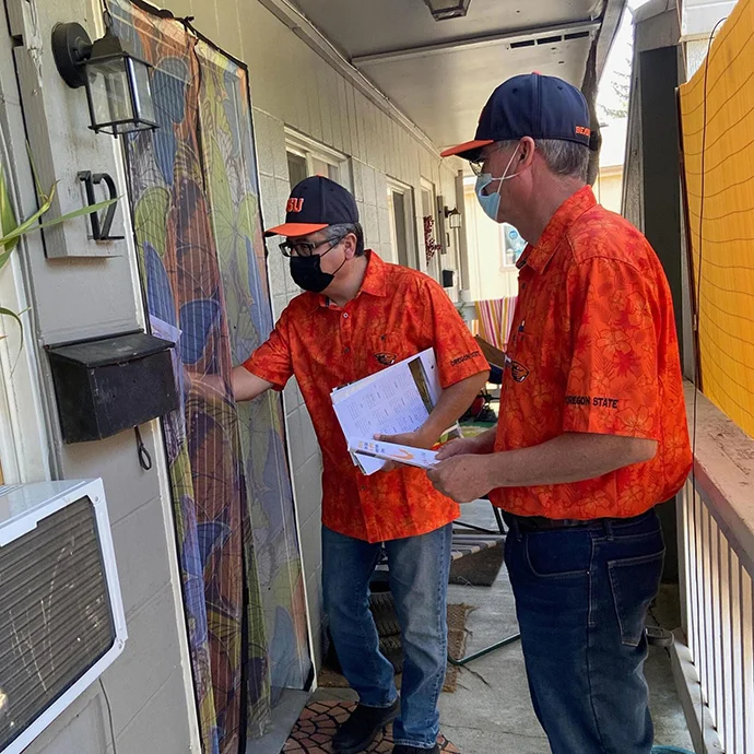 Two men wearing orange Oregon State University (OSU) shirts and face masks stand on a residential porch. One man reaches toward a screen door while holding informational flyers.