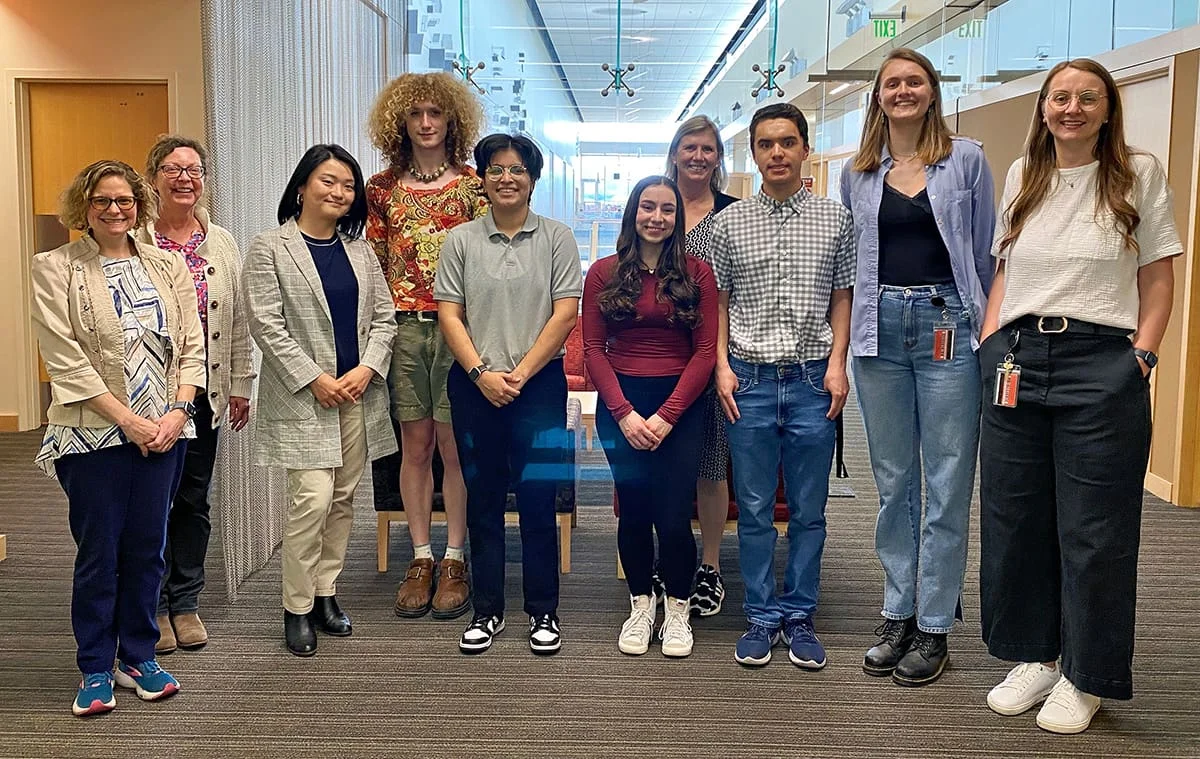 Group of ten people standing in an office setting. They are diverse in age, gender expression, and attire. Background includes doors, glass walls, and ceiling lights.