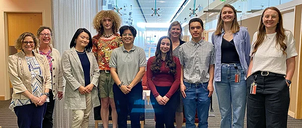 Group of ten people standing in an office setting. They are diverse in age, gender expression, and attire. 
