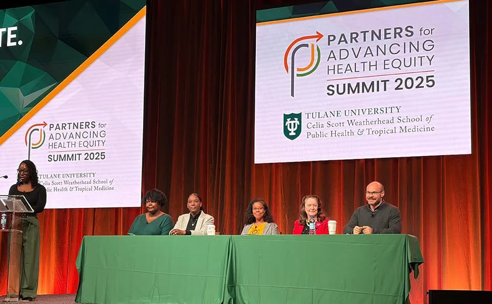 Stage view of the 'Partners for Advancing Health Equity Summit 2025', showing a speaker at the podium, a panel of 5 diverse people behind a table, and the event logo displayed on screens.