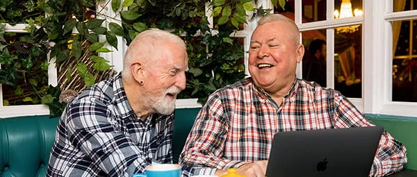 Two older men laugh while looking at a laptop in a cafe.