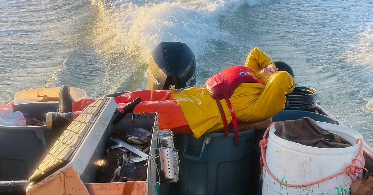 Commercial fisher in a life vest and rain gear takes a break, reclining on the stern of a boat while underway at sea.