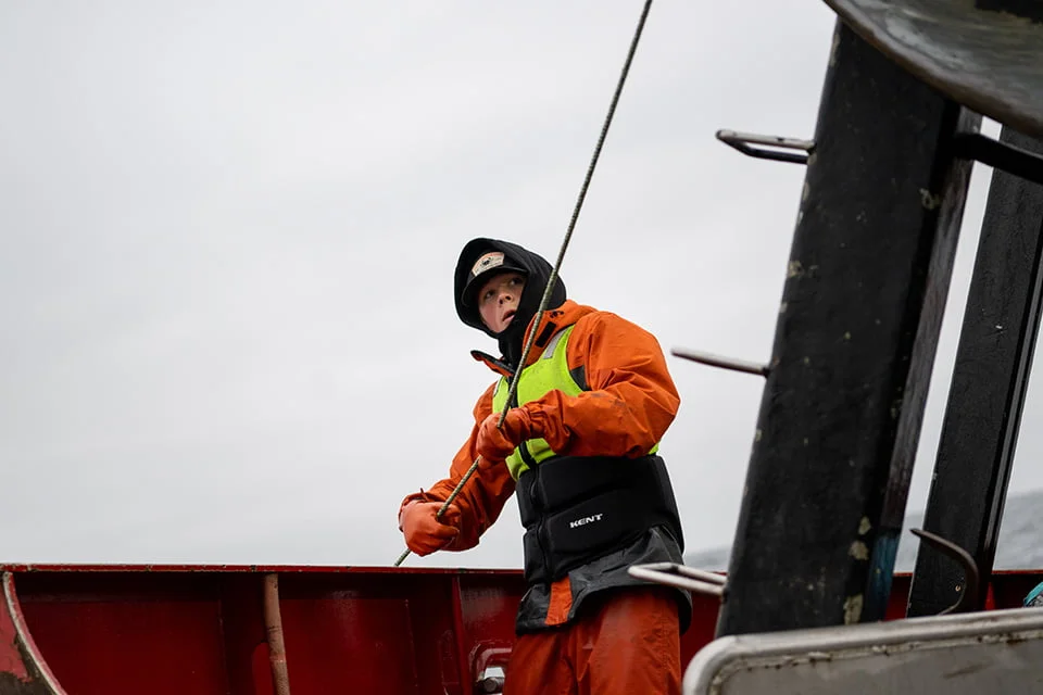 A deckhand in high-visibility safety gear operates lines on a commercial fishing vessel during gray weather conditions.