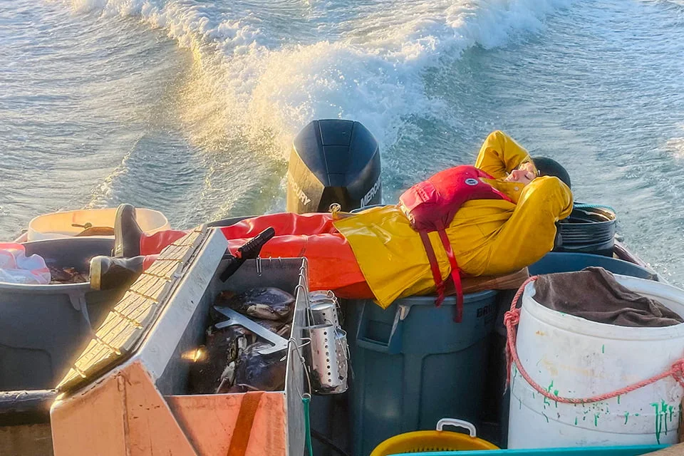 Commercial fisher in a life vest and rain gear takes a break, reclining on the stern of a boat while underway at sea.