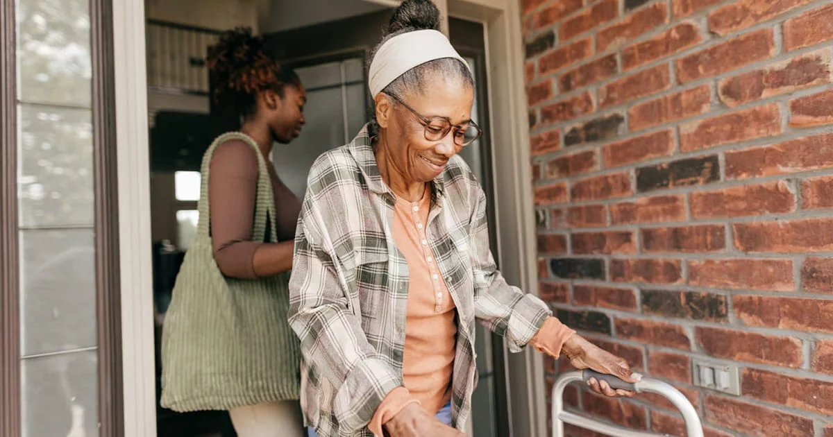 A caregiver helping an elderly woman with a walker leave her home. Emphasizes independence and support.