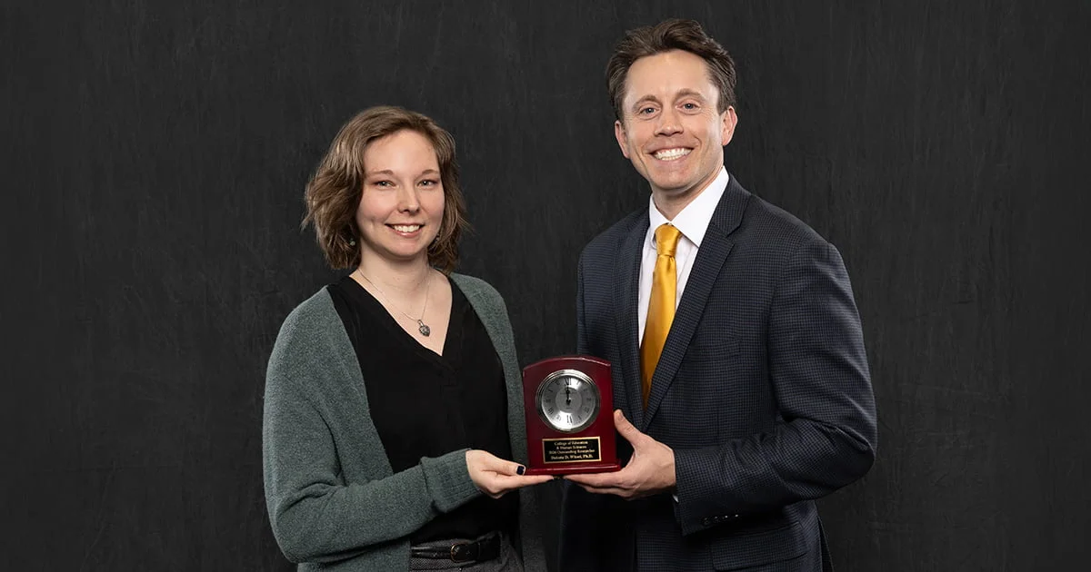 A woman and a man smiling and holding an award clock on a black background.