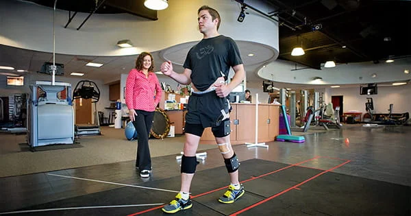 Person wearing knee braces standing on red-lined floor mat in a rehabilitation or fitness center, with another person nearby.