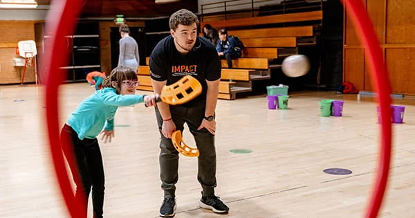 Adaptive PE: Girl using a paddle to hit a ball with assistance from an instructor in a school gymnasium. Promoting inclusivity and physical activity.