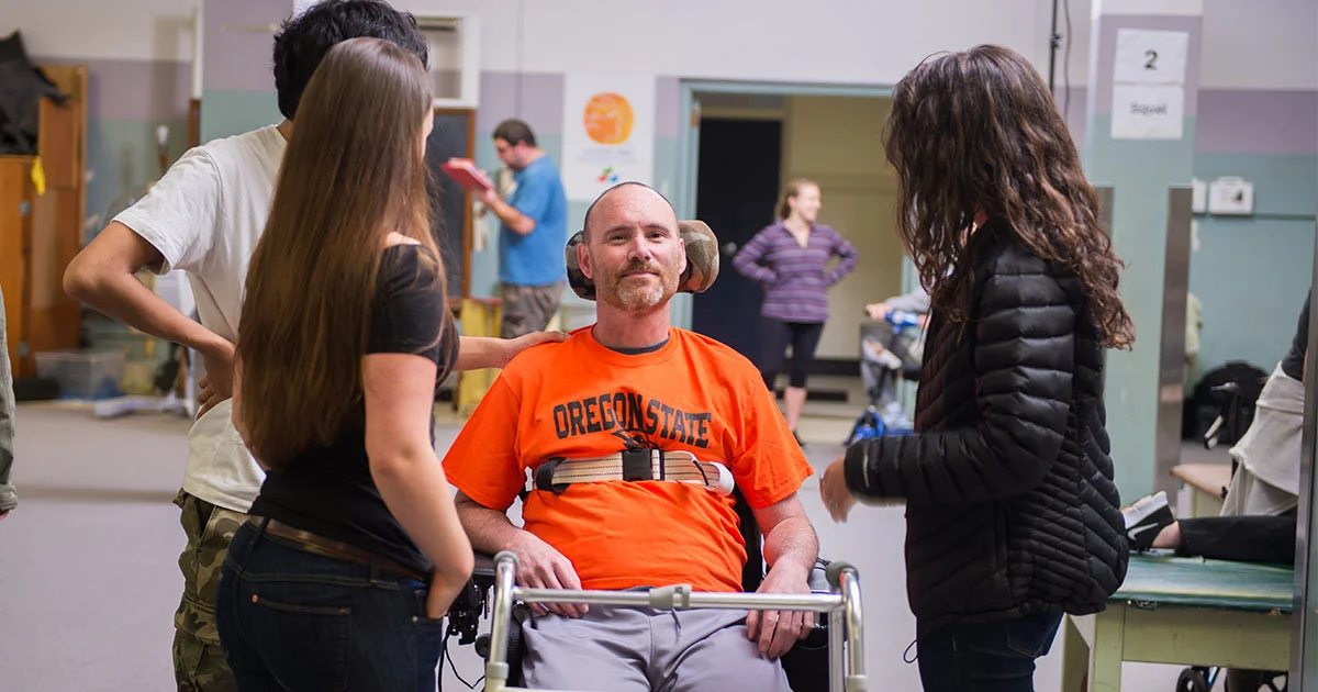 Group interacting with a man in a wheelchair wearing an Oregon State shirt. The setting is a rehabilitation center.