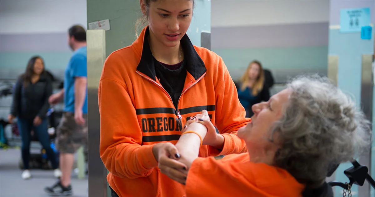Student assisting an older woman in a wheelchair with physical therapy. The student is wearing an Oregon sweatshirt.