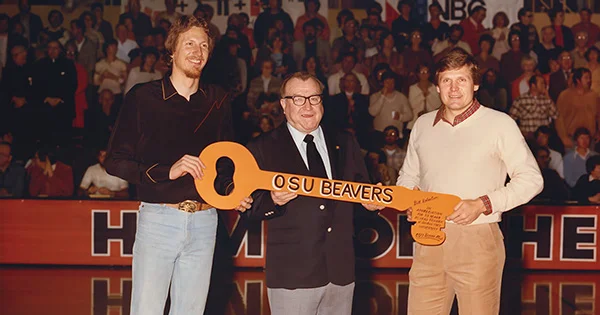 Three men holding a large orange key with 'OSU BEAVERS' written on it, smiling at an event with a crowd in the background.